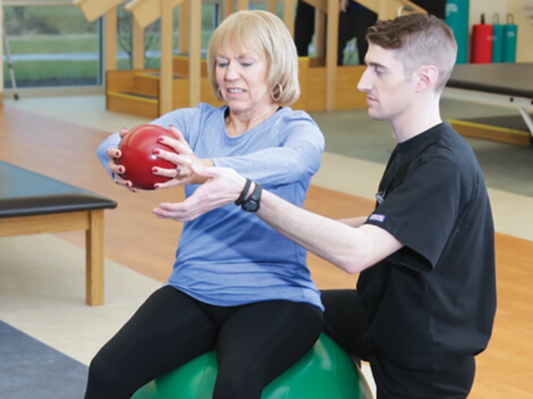 Patient sits on an exercise ball and does arm exercises with physical therapist. 
