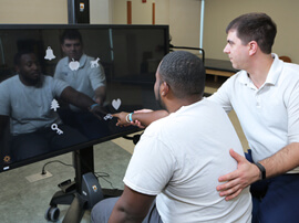 Male therapist works with male patient pointing to shapes on a large TV monitor.