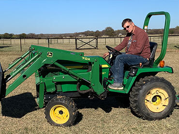 Daniel sitting upon a green and yellow tractor with fence and grassy pasture in background.