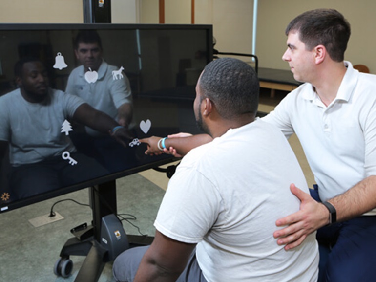 Male therapist works with male patient pointing to shapes on a large TV monitor.