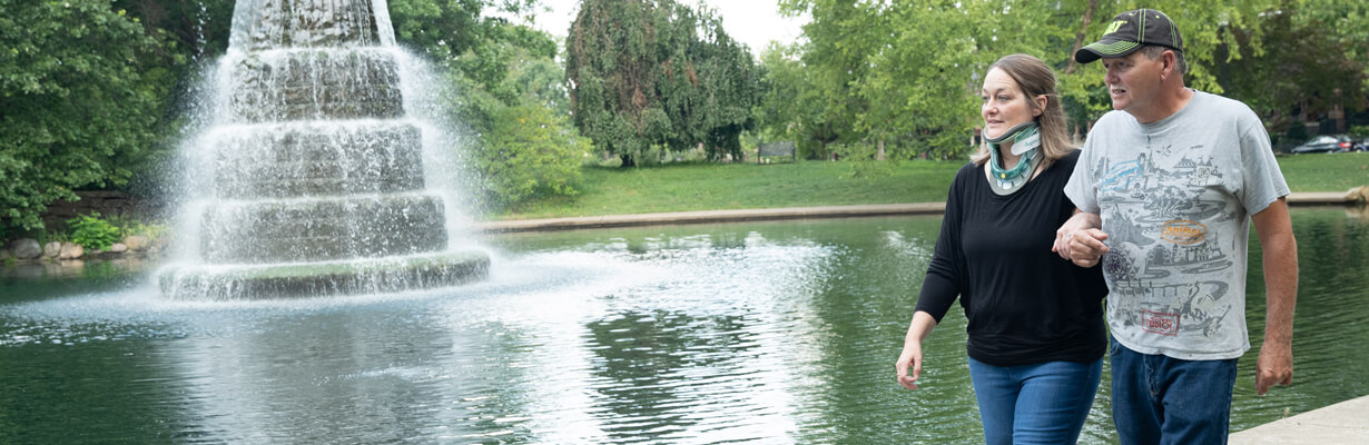 Female patient wearing neck brace walking arm in arm with man in front of fountain.