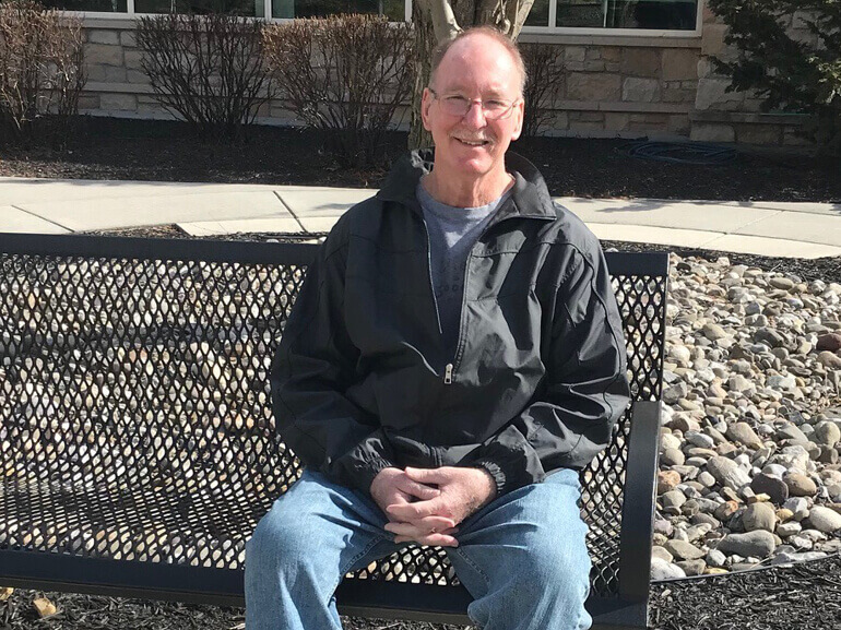 Richard sitting outside on a bench in front of a pebbled landscape and smiling.