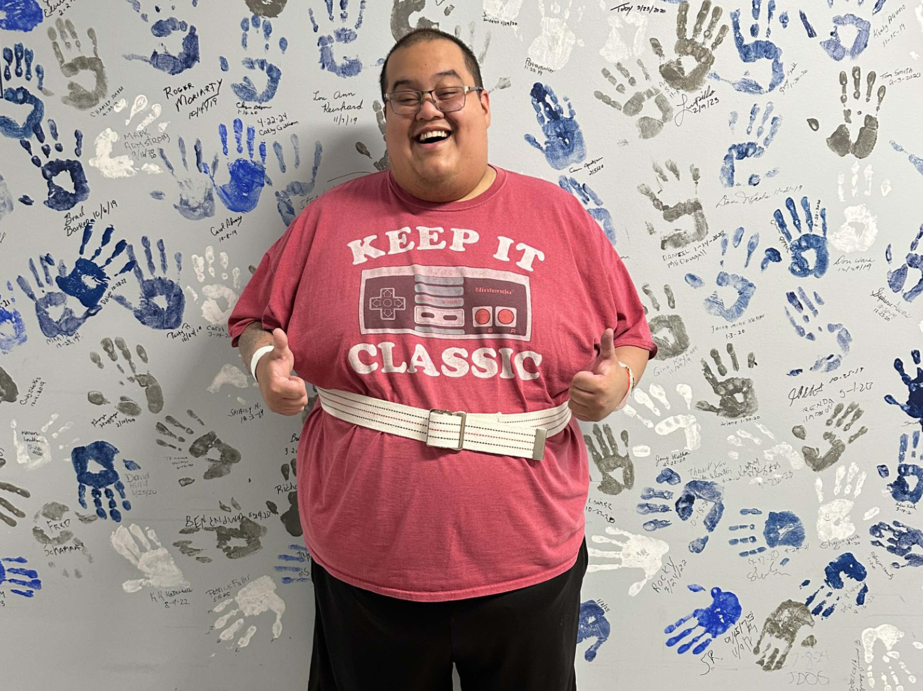 Daniel Santana offers a wide grin and a thumbs up sign as he stands with his back against a wall covered in handprints and signatures. 